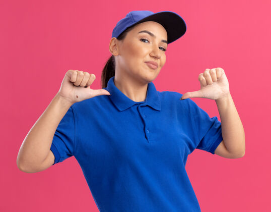拇指身穿蓝色制服 头戴鸭舌帽的年轻女送货员快乐而自信地用拇指指着站在粉色墙上的自己站立指点年轻