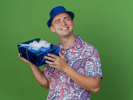 盒子面带微笑的年轻人一边看一边戴着蓝色帽子拿着礼品盒隔离在绿色背景上礼物年轻人小伙子