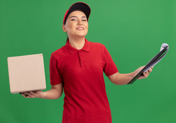 送货身穿红色制服 头戴鸭舌帽的年轻女送货员拿着纸板箱 拿着剪贴板 站在绿色的墙边 面带微笑地看着前面制服剪贴板帽子