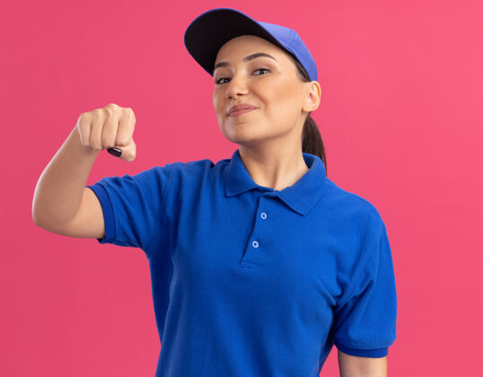 握紧身穿蓝色制服 头戴鸭舌帽的年轻女送货员站在粉红色的墙上 紧握着拳头 自信地微笑着立场年轻显示