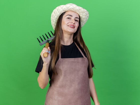 女孩微笑美丽的园丁女孩穿着制服戴着园艺帽子把耙子放在肩膀上隔离在绿色背景上帽子耙子穿