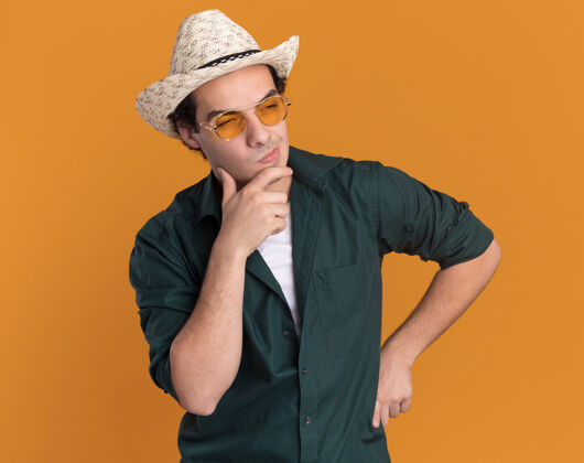 站一个穿着绿衬衫 戴着夏令帽 戴着眼镜的年轻人站在橘色的墙上 茫然地看着一边男人拼图衬衫