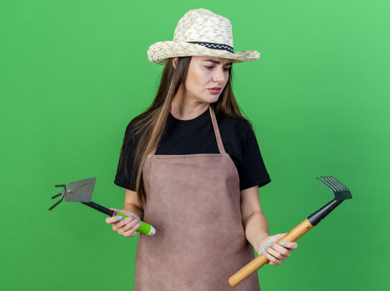 耙子迷茫美丽的园丁女孩穿着制服戴着园艺帽拿着锄头耙子看着耙子在她的手上孤立的绿色背景园艺帽子持有