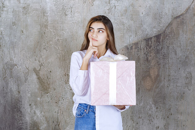 思考一个穿着白衬衫的女人手里拿着一个用白丝带包着的粉色礼盒 看上去困惑和犹豫不确定生日年轻人