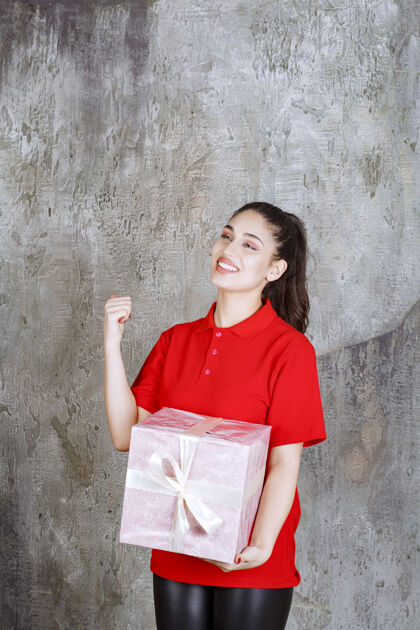 人年轻女子手持一个用白丝带包裹的粉红色礼盒 并显示积极的手势力量女人享受