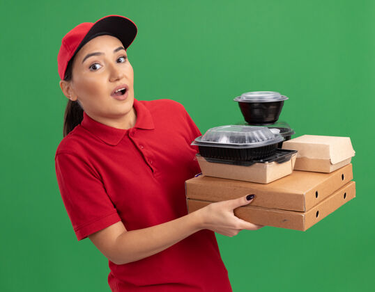 送货身穿红色制服 头戴鸭舌帽 拿着比萨饼盒和食品包的年轻送货员站在绿色的墙壁上 困惑和担忧地看着前方制服帽子盒子