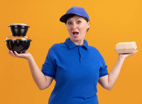 站着身穿蓝色制服 头戴鸭舌帽的中年女送货员站在橘色的墙上惊讶地看着前面的食品包送货拿着帽子