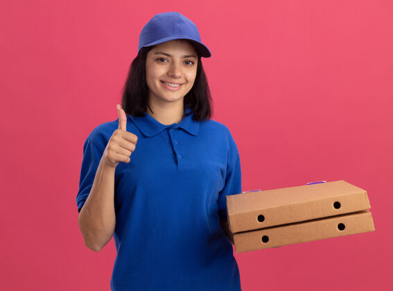 披萨身穿蓝色制服 头戴鸭舌帽 手持披萨盒的年轻送货女孩站在粉红色的墙上 脸上露出幸福的笑容 竖起大拇指年轻微笑盒子