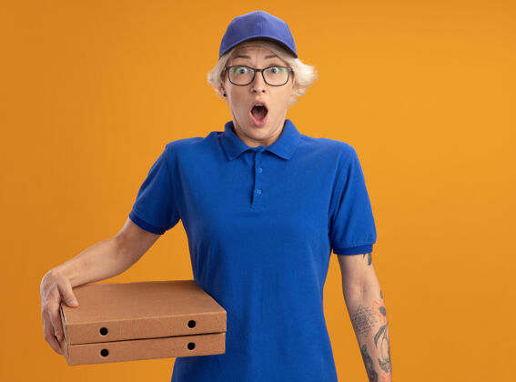 穿身着蓝色制服 戴着眼镜 拿着比萨饼盒的年轻送货员在橙色的墙上惊讶不已帽子惊喜年轻人