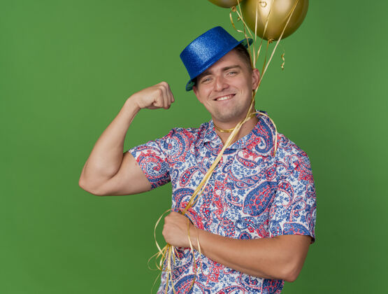 脖子带着微笑的年轻人戴着蓝色帽子拿着气球绑在脖子上孤立的绿色背景抱着戴着微笑