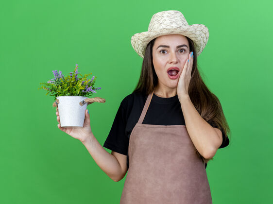 惊喜一个穿着制服的美丽的园丁女孩戴着园艺帽 手里拿着花盆里的花 手放在绿色背景上的脸颊上女孩穿制服