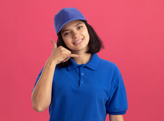 手势穿着蓝色制服 戴着帽子的年轻送货女孩站在粉红色的墙上微笑着打电话给我女孩做送货