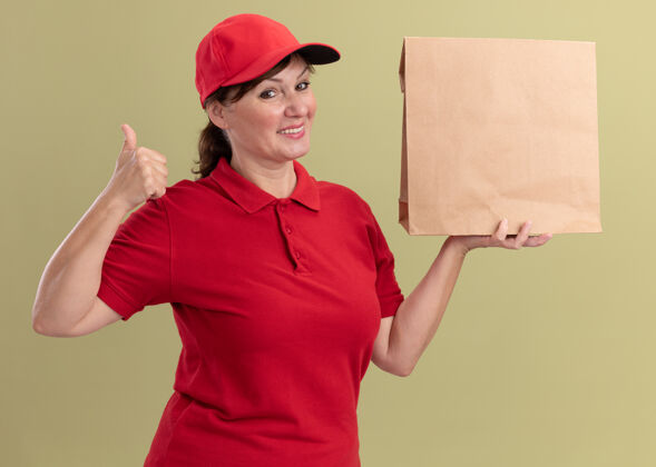 女人快乐的中年女送货员穿着红色制服 戴着帽子 拿着纸包 微笑着自信地站在绿色的墙上竖起大拇指 看着前面中间拇指自信
