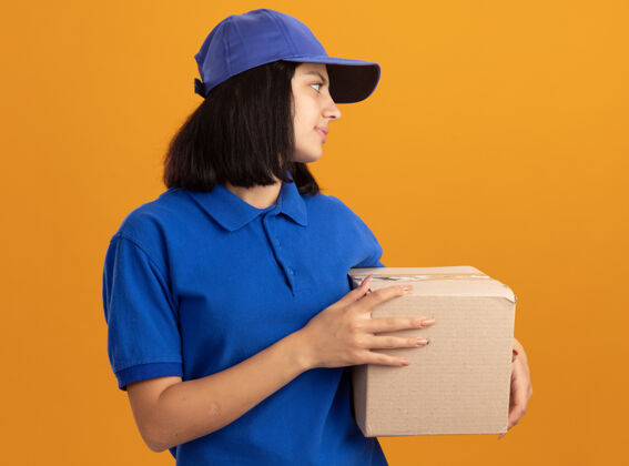 抱着身穿蓝色制服 戴着帽子的年轻送货员站在橙色的墙上 友好地微笑着站在一旁看着纸箱站着朋友送货