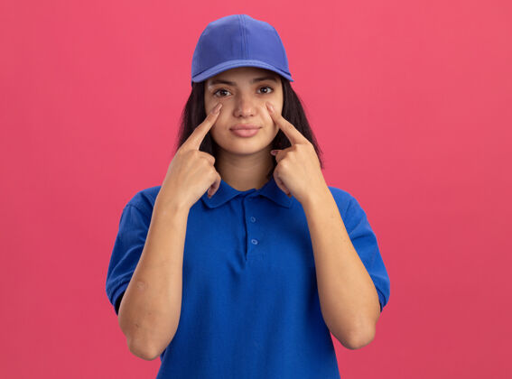 手指穿着蓝色制服 戴着帽子的年轻送货女孩站在粉红色的墙上用食指指着她的眼睛帽子目录女孩