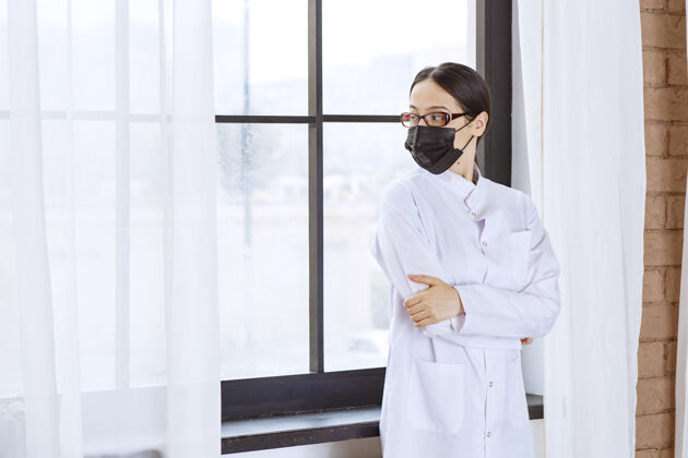 从业者戴着黑面具和眼镜的医生站在窗边护理模特治疗