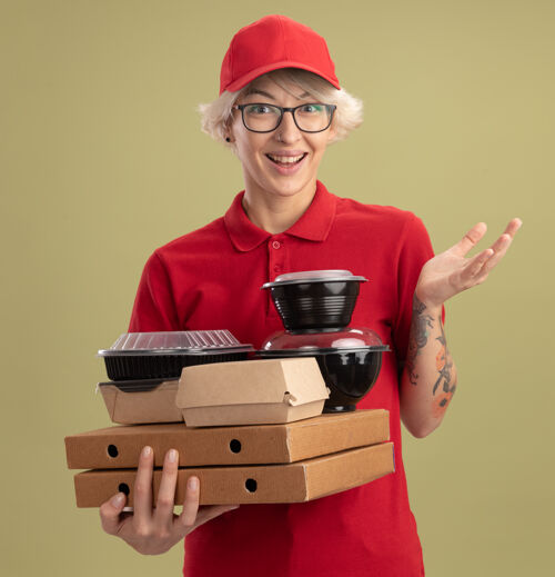披萨身着红色制服 戴着眼镜 拿着比萨饼盒和食品包 微笑着站在绿色墙壁上的快乐的年轻女送货员盒子拿着制服