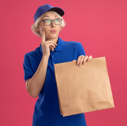 帽子身穿蓝色制服 戴着眼镜 拿着纸包的年轻女送货员隔着粉色的墙 疑惑地看着一旁送货靠边站眼镜