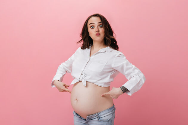 深色蓝眼睛的黑发女人穿着白色不规则剪裁的衬衫和牛仔裤做滑稽的脸怀孕的女人指着她的腹部在粉红色的背景怀孕室内为人父母