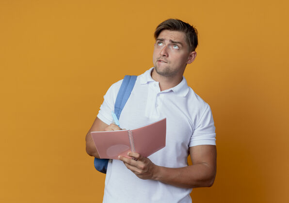 包抬头望着迷茫的年轻帅哥 背着书包 拿着笔记本和笔 孤立在橙色的背景上学生背笔记本