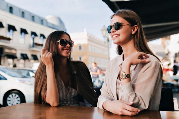年轻两个戴着墨镜的开朗女孩在广场上愉快地交谈着 带着完美的笑容戴着墨镜女士黑发城市