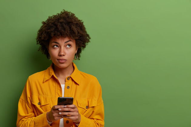 手机沉思的女人在智能手机上使用现代应用 思考信息内容 专注于上面 与现代科技保持联系 穿着时尚的衣服在绿色墙壁的空白处短信现代发型