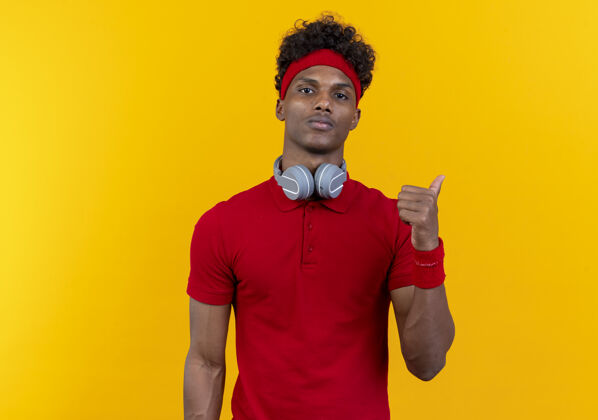 耳机看着镜头 年轻的美国黑人运动型男子戴着头带和腕带 戴着耳机 指着隔离在黄色背景上的一边 留着复印空间运动非裔美国人壁板