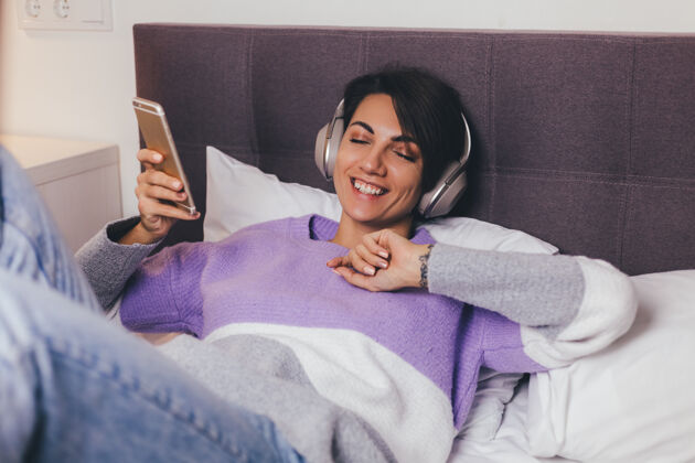 床快乐的女人在家舒适的床上穿着暖和的衣服套头衫 听音乐耳机科技休息