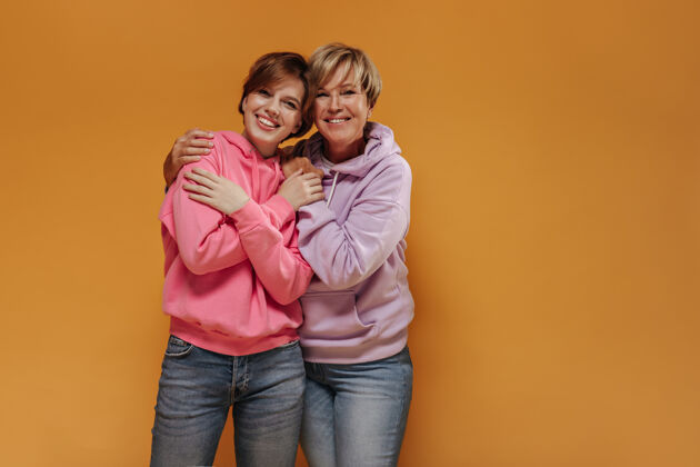 休闲迷人的两位女士 带着可爱的笑容 留着一头酷酷的短发 穿着时髦的粉色运动衫和牛仔裤 在与世隔绝的背景下拥抱在一起衬衫粉色中等