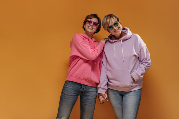 女儿漂亮时尚的两位女士 戴着凉爽的太阳镜和粉色连帽衫 在橙色孤立的背景下微笑着 手牵着手两个年龄休闲