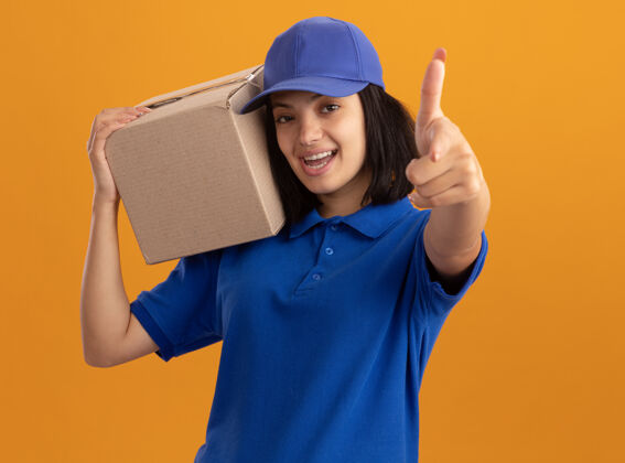 帽子身穿蓝色制服 戴着帽子的年轻送货员拿着纸板箱微笑着 食指指着橙色的墙上年轻举行目录