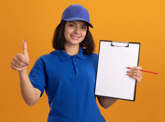 显示身穿蓝色制服 戴着帽子的年轻送货女孩站在橙色的墙上 手里拿着空白的纸和铅笔 微笑着竖起大拇指页面铅笔空白