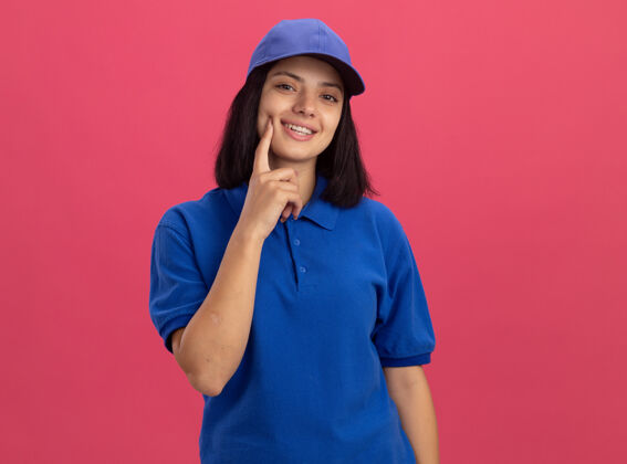 年轻身穿蓝色制服 头戴鸭舌帽的年轻送货员站在粉红色的墙上 脸上露出幸福的笑容 手指放在脸颊上手指交货脸颊