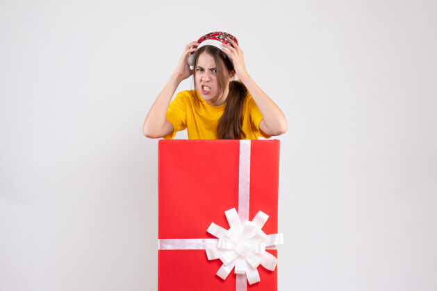 包前视图愤怒的女孩 戴着圣诞帽站在圣诞礼物后面圣诞节人购物