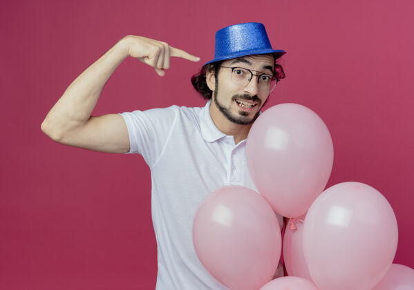 眼镜高兴的帅哥戴着眼镜 戴着蓝色的帽子 手里拿着气球 在一旁点着粉红色的背景隔离请戴帅气手持