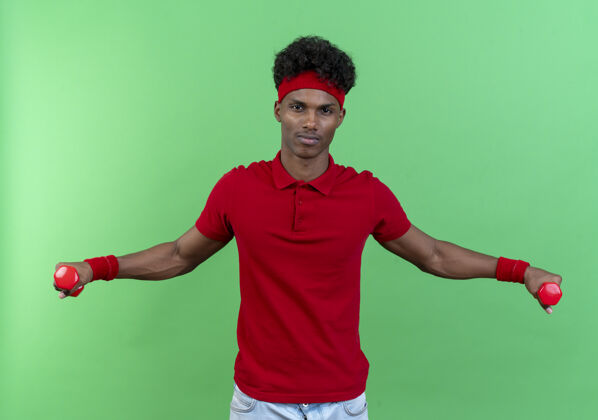 运动看着镜头年轻的黑人运动男子戴着头带和腕带锻炼哑铃隔离在绿色背景年轻穿着头带
