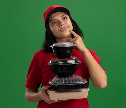 持有快乐的年轻送货女孩穿着红色制服 戴着帽子 手里拿着一叠食品包 站在绿色的墙边茫然地看着一边一旁立场堆栈