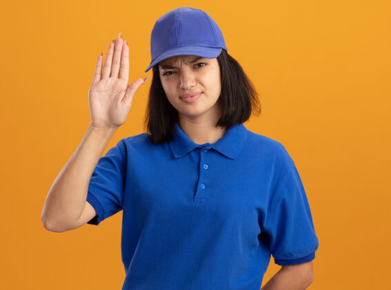 立场身穿蓝色制服 头戴鸭舌帽的年轻送货女孩 严肃的脸上露出张开的手站在橙色的墙上脸制服交货