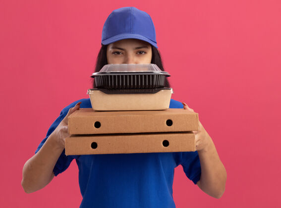 食物身穿蓝色制服 头戴鸭舌帽 拿着披萨盒和食品包的年轻送货女孩站在粉红色的墙上 表情严肃年轻脸送货