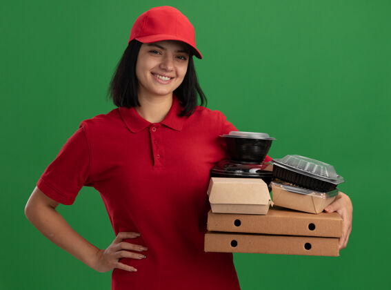 盒子身着红色制服 头戴鸭舌帽 手持披萨盒和食品包的快乐年轻送货女孩站在绿色的墙上 开心地微笑着站着女孩年轻人