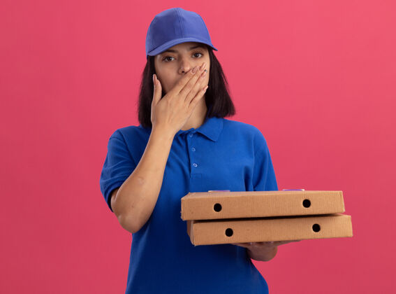 女孩身穿蓝色制服的年轻送货女孩拿着比萨饼盒 手捂着嘴站在粉红色的墙上 震惊不已制服盒子手