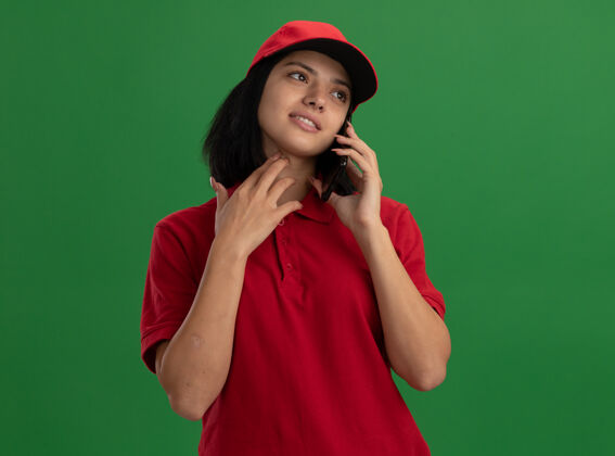微笑身穿红色制服 头戴鸭舌帽的年轻送货员站在绿色的墙上 一边微笑着讲着手机帽子而手机