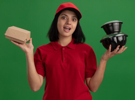 制服身穿红色制服 头戴鸭舌帽 拿着食品包的年轻送货女孩站在绿色的墙上 又高兴又惊讶食物送货站着
