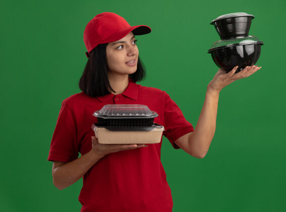 年轻身穿红色制服 头戴鸭舌帽 拿着食品包的年轻送货女孩站在绿色的墙上 看上去自信满面笑容食物盒子站着