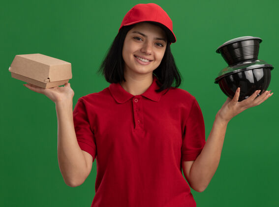 食物身着红色制服 头戴鸭舌帽 手持食品包装 微笑友好地站在绿色墙壁上的快乐少女年轻制服送货