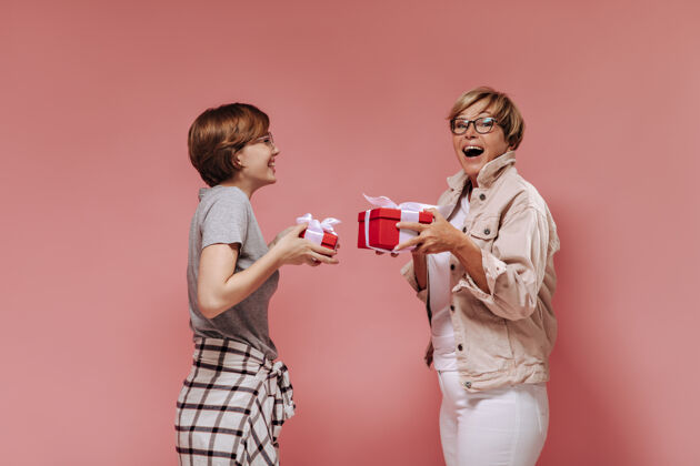 短发欢快的两位女士 留着现代短发 穿着时髦的衣服 戴着眼镜 手里拿着红色的礼盒 在粉色的背景下欢呼雀跃中等灰色夹克