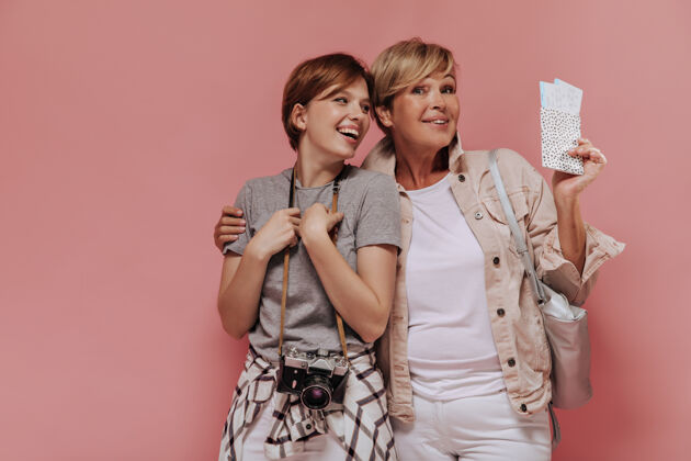 短发漂亮的两个女人 时尚的短发 穿着时髦的衣服 拥抱着 笑着 拿着两张票和相机在粉红色的背景上成人休闲夹克