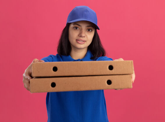 披萨身穿蓝色制服 头戴鸭舌帽 手持披萨盒的年轻送货员站在粉色的墙上 显得自信满满制服拿着站着