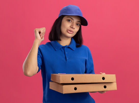 站着身穿蓝色制服 头戴鸭舌帽 手持披萨盒 握紧拳头的年轻送货女孩站在粉色的墙上 看上去很自信年轻女孩帽子