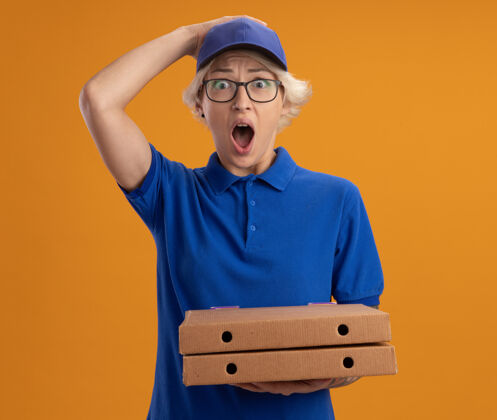 惊喜身着蓝色制服 戴着眼镜 拿着比萨饼盒的年轻送货员在橙色的墙上惊讶不已眼镜穿送货